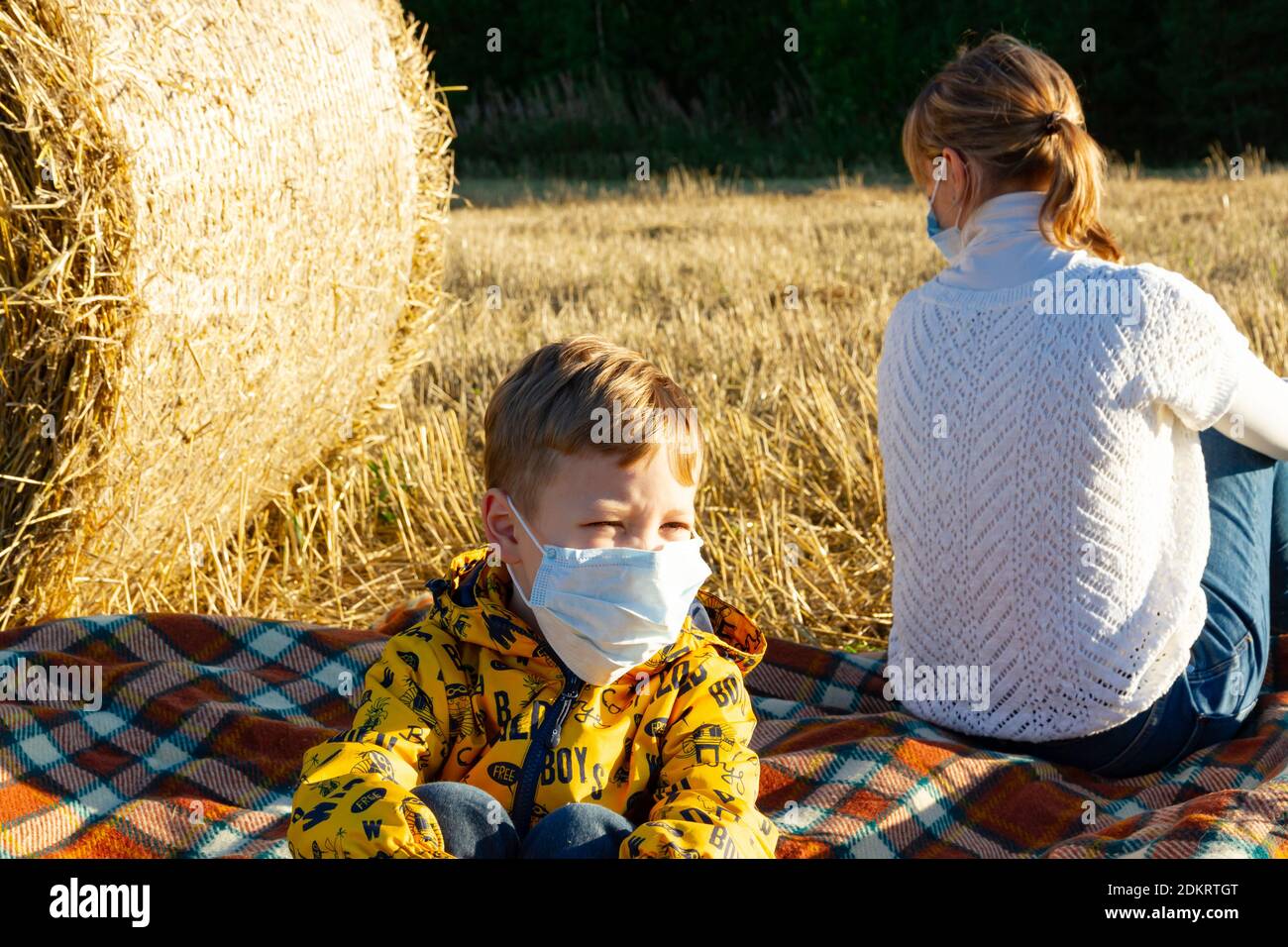 mamá y el bebé se sientan en el suelo en la naturaleza el fondo de los pajar en máscaras médicas Foto de stock