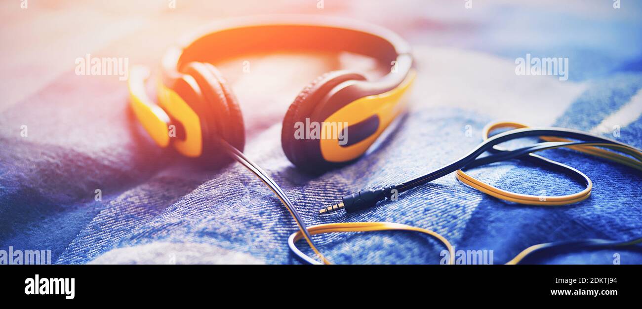 Los grandes auriculares amarillos con un cable largo se encuentran sobre  una manta de lana de cuadros azul suave, iluminada por la luz del sol  brillante. Inspiración y aficiones. Escucha a ple