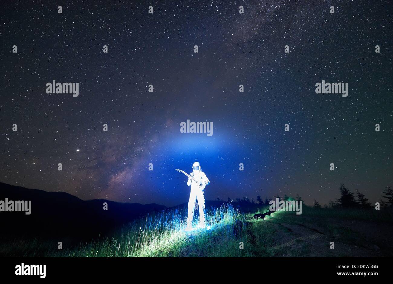 Cosmonauta iluminado en traje blanco y casco de pie en medio de la pradera  de montaña sosteniendo la guitarra, increíble cielo nocturno lleno de  estrellas en el fondo. Concepto de viaje espacial