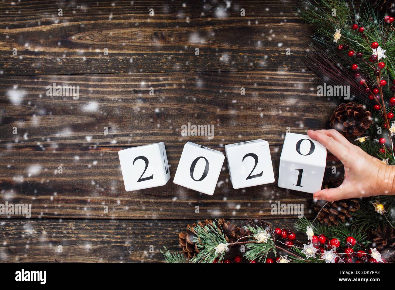 La mano de la mujer volteando los 2021 bloques de calendario de madera de año Nuevo a 2020. Luces de árbol de Navidad, ramas de pino, bayas rojas de invierno y nieve. Vista superior. Foto de stock