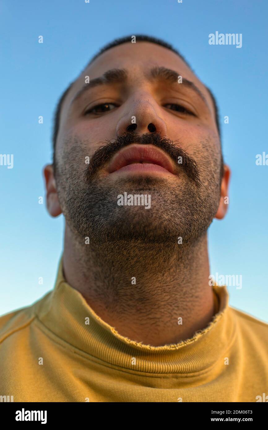 Retrato de un joven con bigote mirando hacia abajo a la cámara delante del cielo azul con un puente amarillo Foto de stock