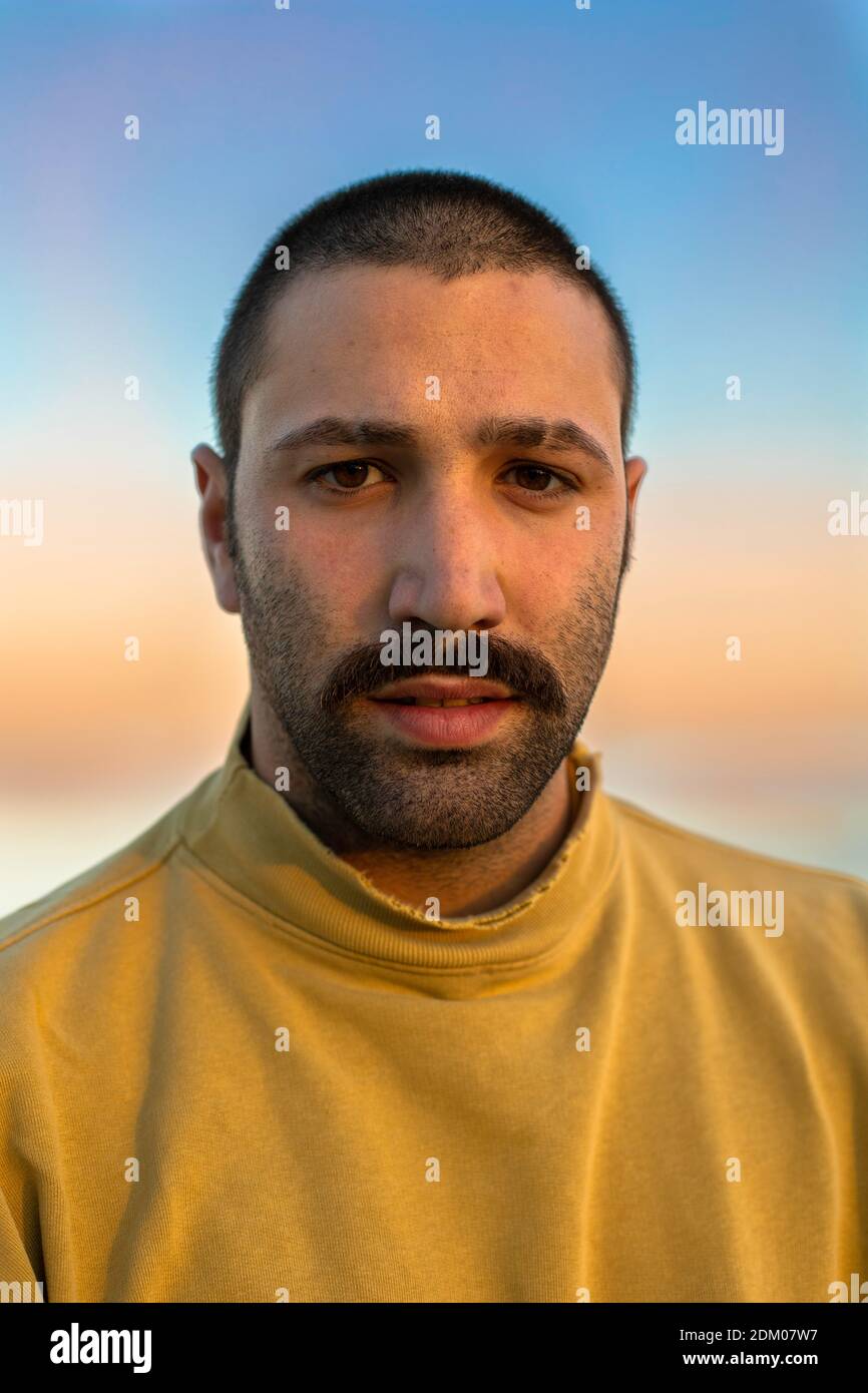 Retrato de un joven con bigote mirando hacia dentro de la cámara frente del cielo al atardecer con un puente amarillo Foto de stock