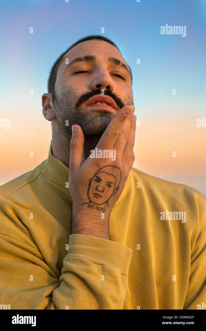 Retrato de un joven con bigote, mano tatuada, cara de ensueño y ojos cerrados frente al cielo del atardecer con un puente amarillo Foto de stock