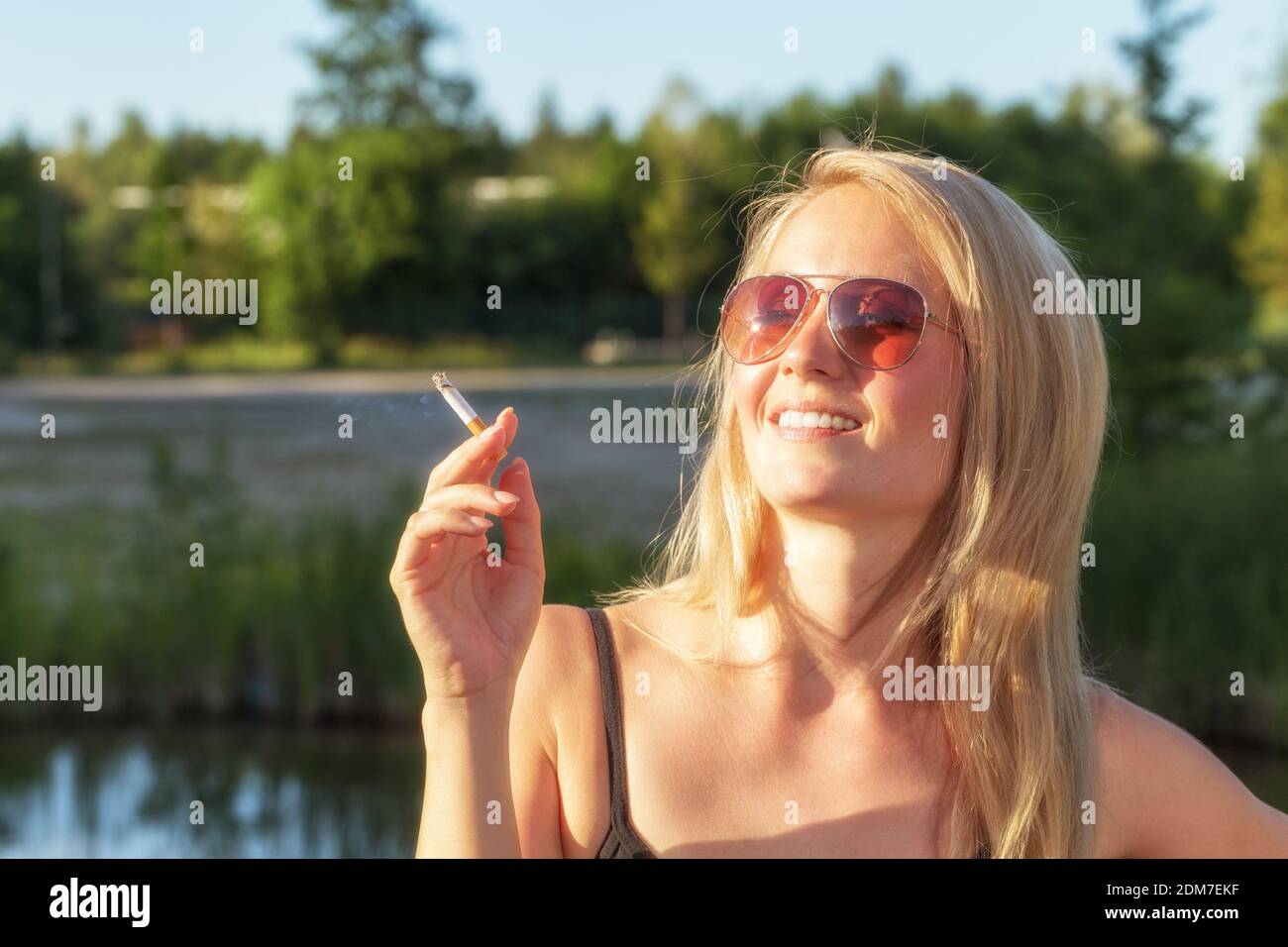 Mujer de adulto medio usando gafas de sol Fumando cigarrillo mientras se encuentra de pie contra los árboles en el parque Foto de stock