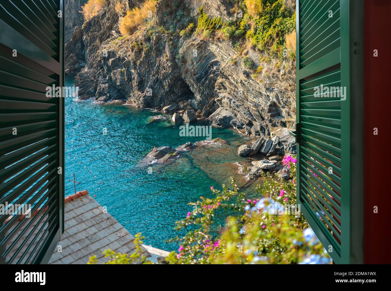 Vista desde una ventana abierta sobre una pequeña bahía con nadadores sentados en las rocas, cerca del pueblo de Vernazza, Italia, parte de Cinque Terre Foto de stock
