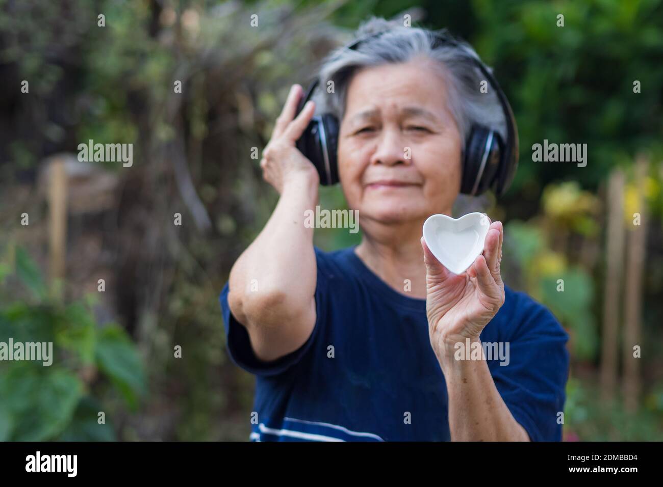 Enfoque selectivo una cerámica blanca en forma de corazón en las manos de  una anciana que lleva puesto unos auriculares inalámbricos escuchando una  canción favorita en el jardín. Día de San Valentín.