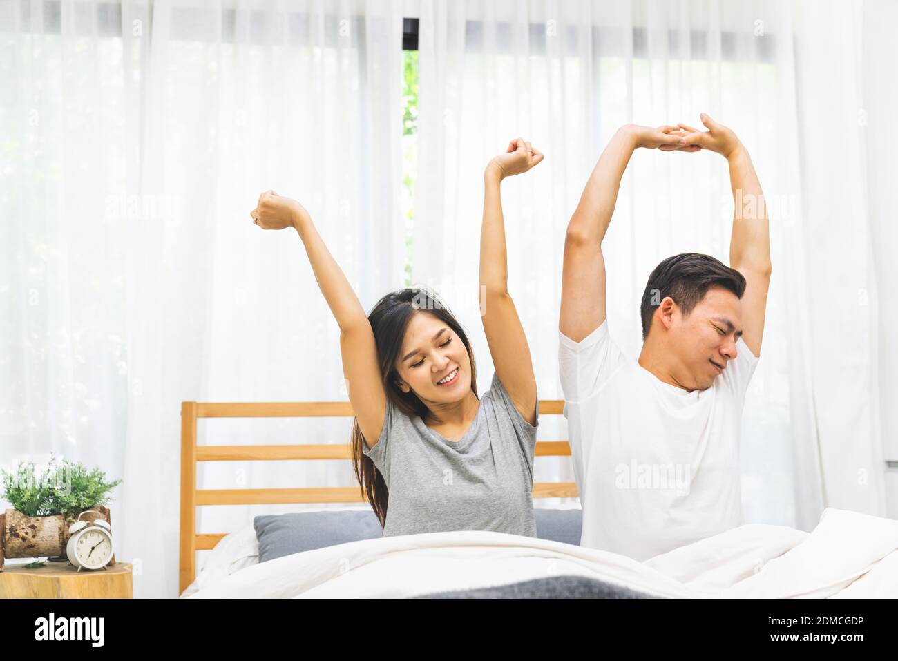 Una pareja joven asiática casada se despierta estirándose juntos en la cama  en el dormitorio. Amanecer y brillo, día perezoso, estilo de vida  doméstico, o concepto de relación de amor para adultos
