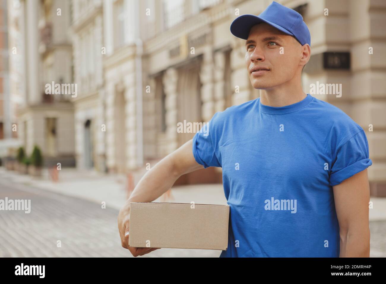 Hermoso mensajero joven en uniforme azul vagando por las calles de la  ciudad con un paquete para cliente, espacio de copia. El hombre de la  entrega que lleva la caja de cartón