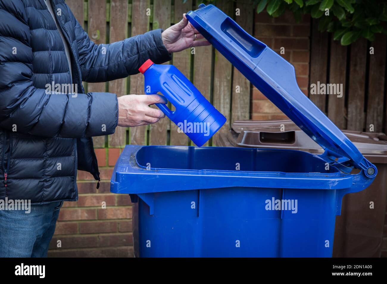 Un hombre responsable colocando una botella de plástico azul en un contenedor de reciclaje con borde de la acera listo para su recogida y reciclaje por el Consejo en el Reino Unido Foto de stock