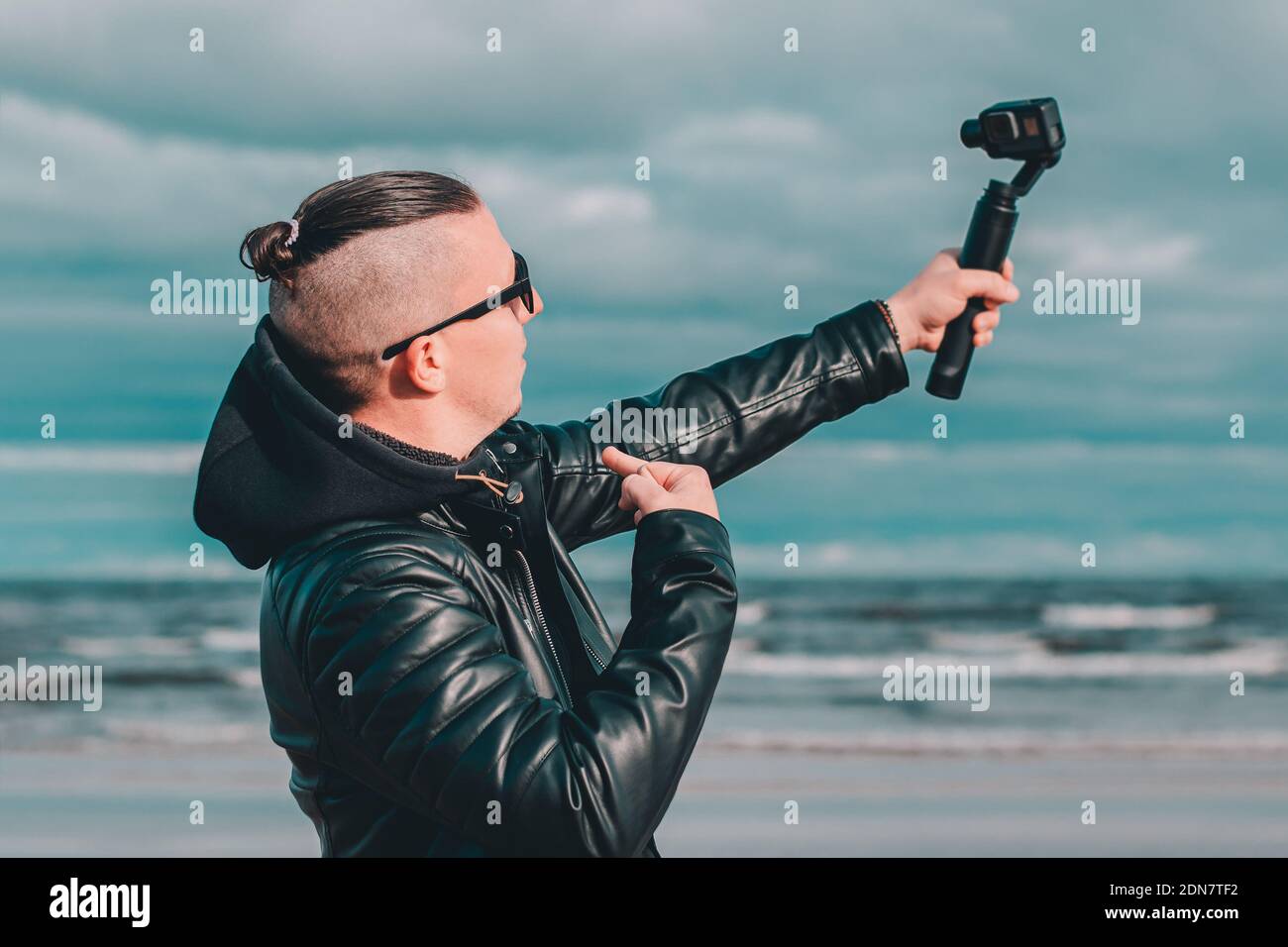 Joven Blogger en gafas de sol haciendo Selfie o streaming de vídeo en la  playa utilizando la cámara de acción con el estabilizador de cámara Gimbal.  Hipster en ropa negra haciendo foto
