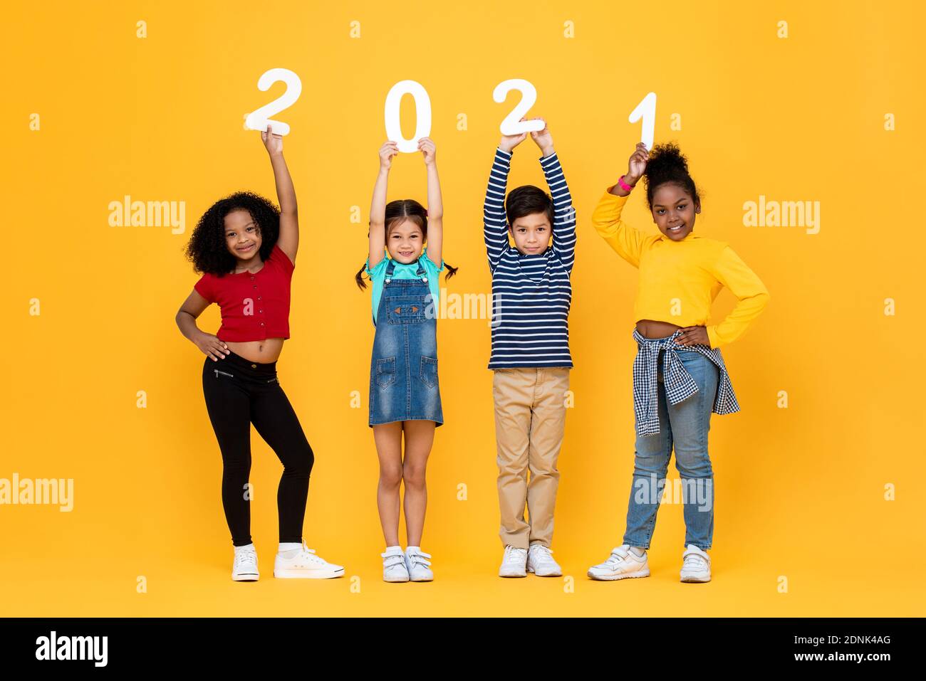Lindos hijos de carreras mixtas sonriendo y sosteniendo 2021 números aislados sobre fondo amarillo para conceptos de año nuevo Foto de stock