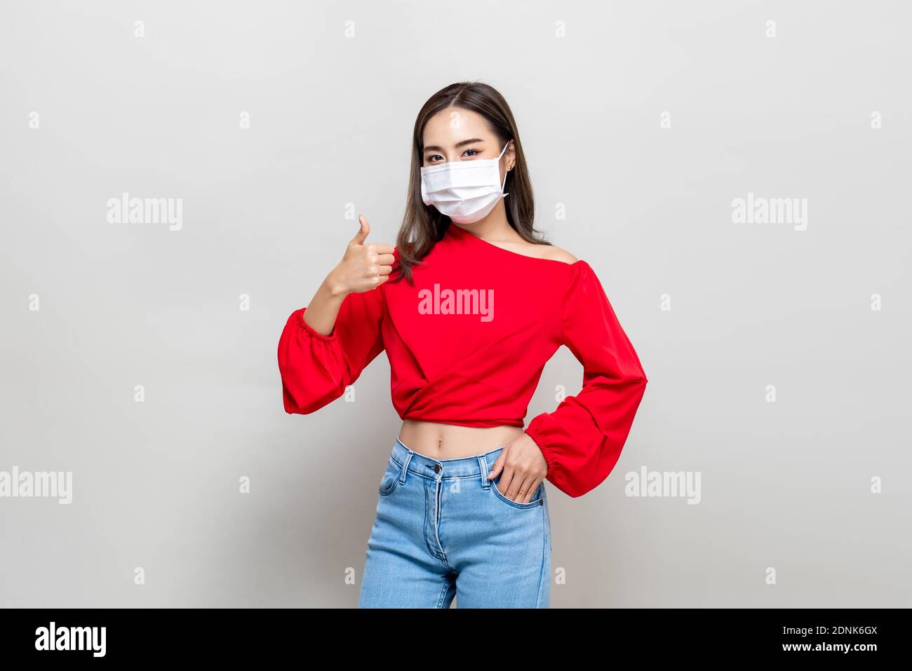 Mujer asiática joven con máscara médica y dando pulgares para arriba sobre fondo de estudio gris claro Foto de stock