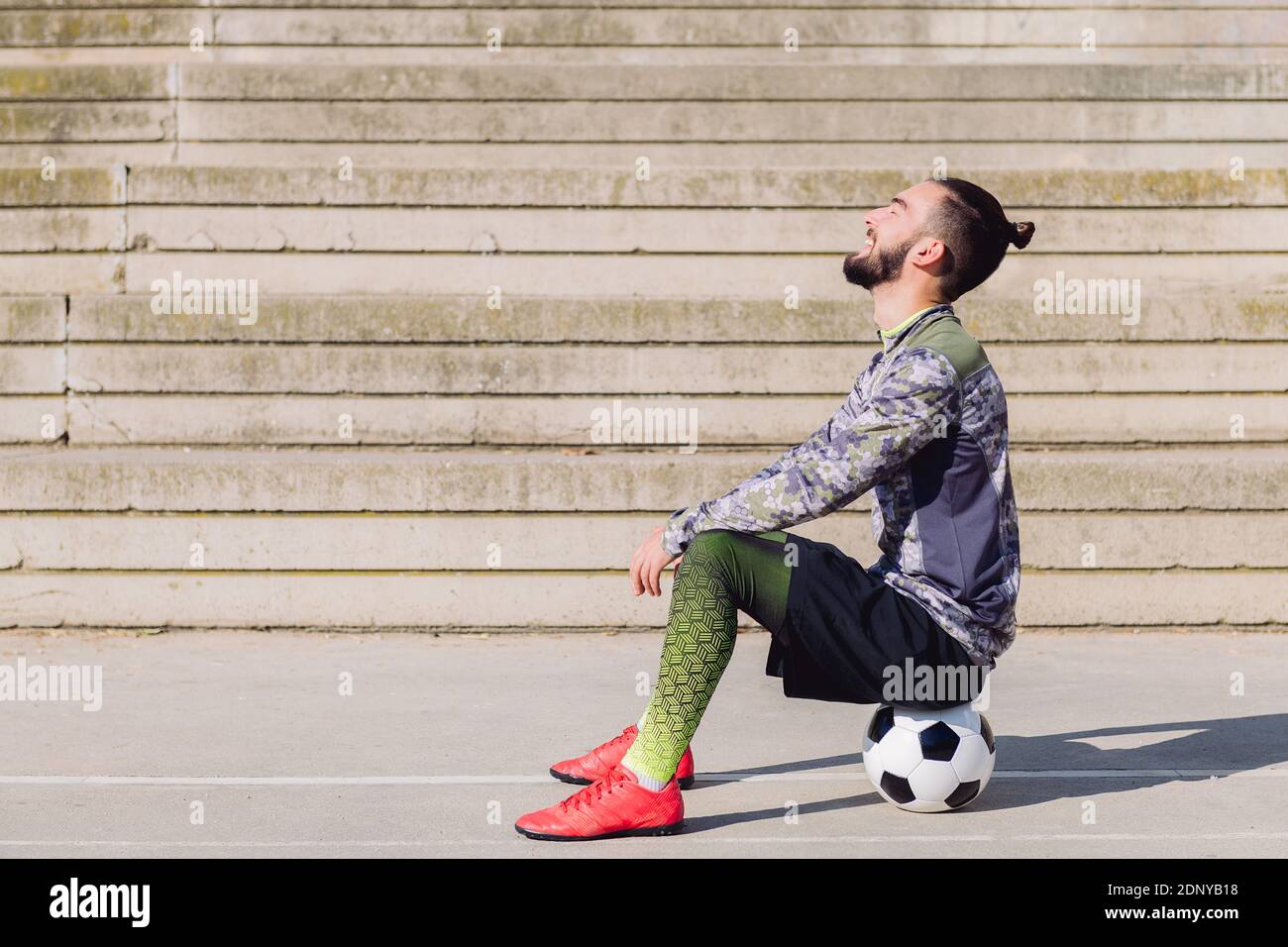 deportista guapo con pelo en un juego de reírse en una cancha de fútbol de  concreto sentado en su balón de fútbol, concepto de estilo de vida  saludable y deporte urbano i