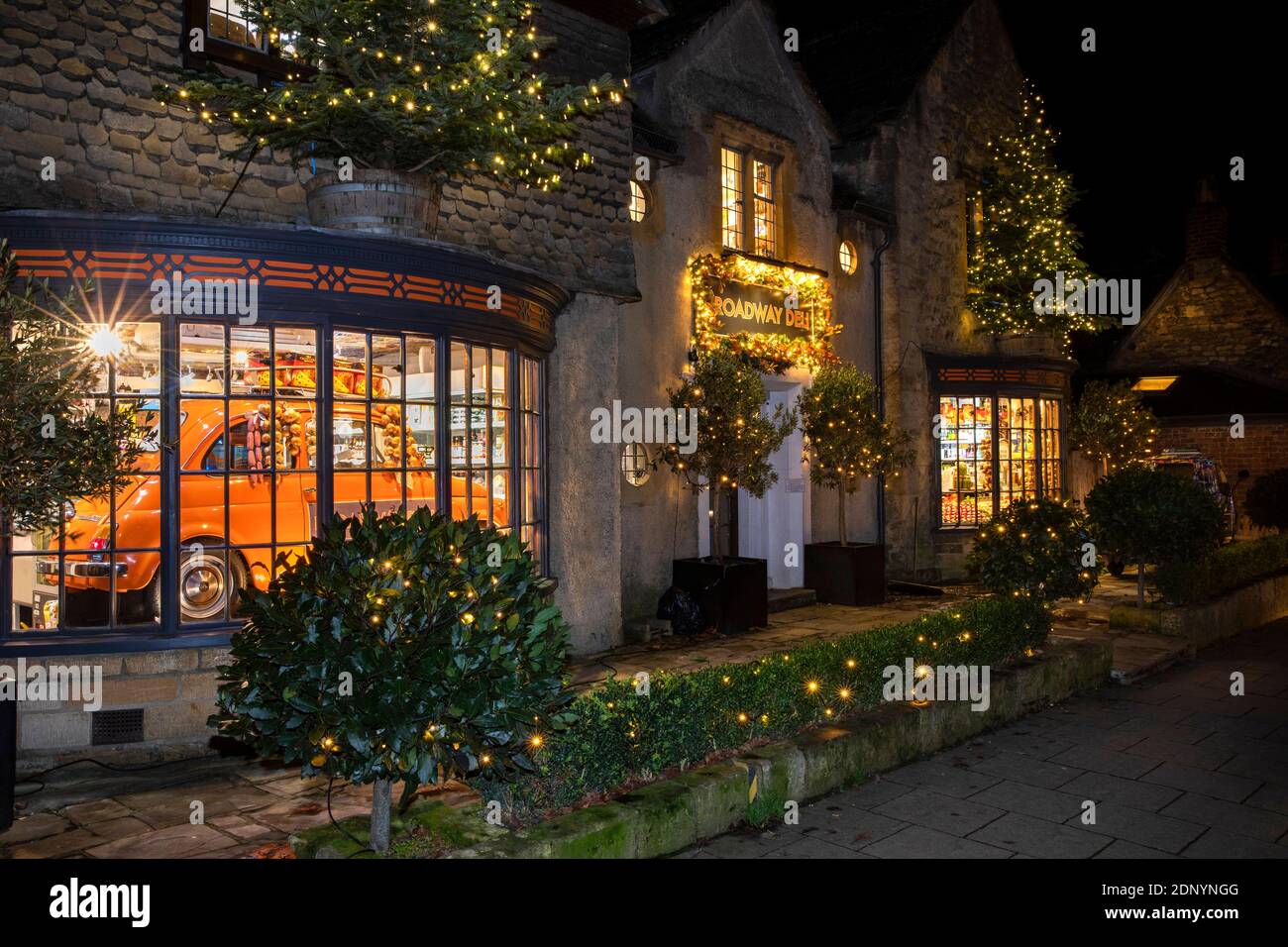 Reino Unido, Gloucestershire, Broadway, High Street, Broadway Deli iluminado para Navidad, con coche en la ventana de la tienda Foto de stock