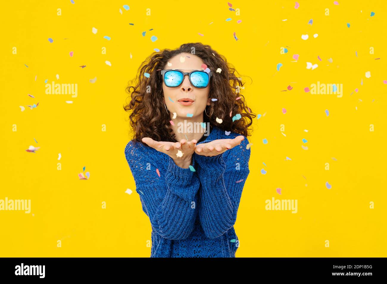 Retrato de una joven con gafas de sol espejadas que soplan confetti el aire frente al fondo amarillo Foto de stock