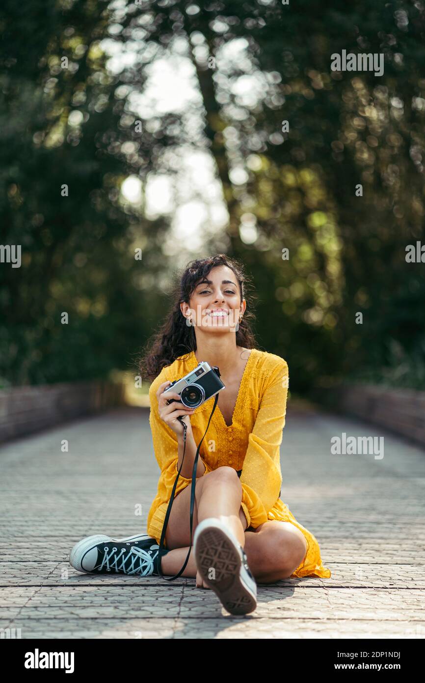 Joven fotógrafo vistiendo vestido amarillo y negro zapatillas sosteniendo una cámara analógica en el paseo marítimo de madera Foto de stock