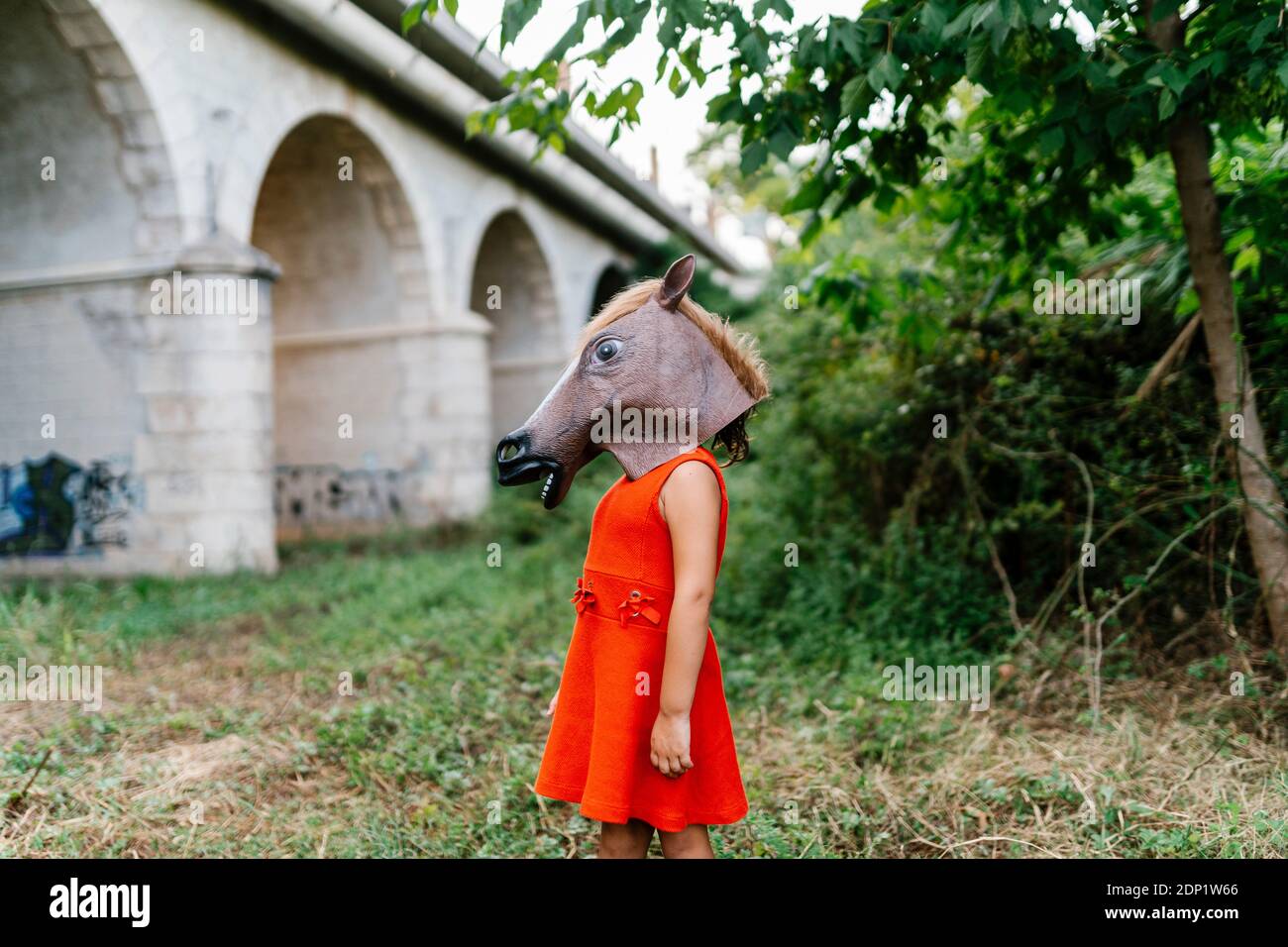 Niña con cabeza de caballo y vestido rojo de pie en la naturaleza bajo un puente Foto de stock