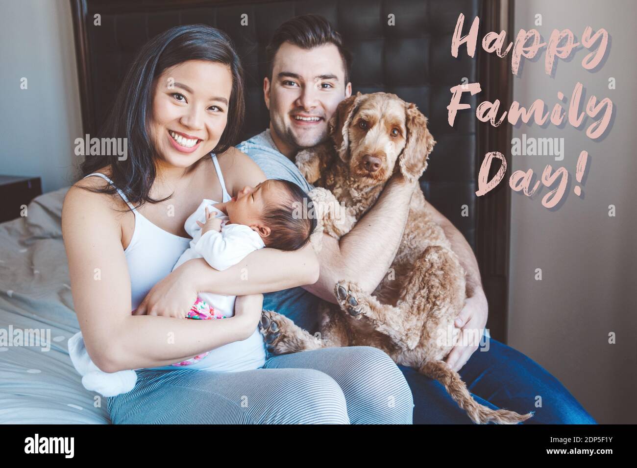 Feliz día de la familia. Tarjeta de vacaciones con texto de saludo. Madre  China asiática sonriente y padre caucásico con bebé recién nacido y perro  mascota. Estilo de vida en casa Fotografía