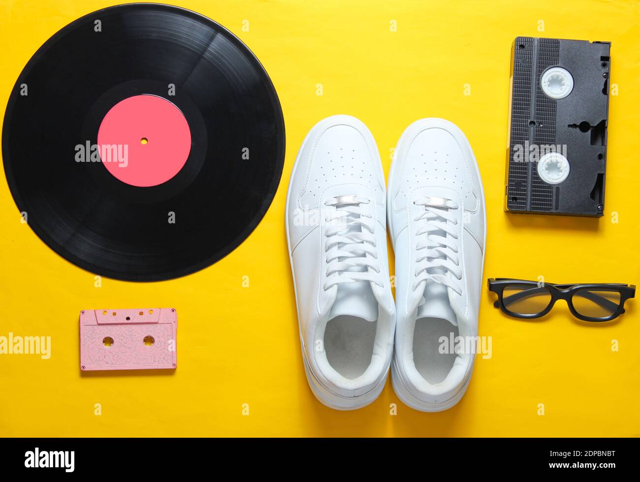 Cultura pop. Zapatillas de lona blancas de cintura baja, placa de vinilo, casete de audio y vídeo, gafas 3d sobre fondo amarillo. Estilo retro. Vista superior, minimalismo Foto de stock