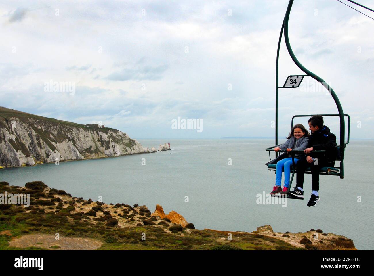Dos jóvenes disfrutando de un paseo en telesilla, Alum Bay, Isla de Wight con el faro de Needles en la distancia Foto de stock