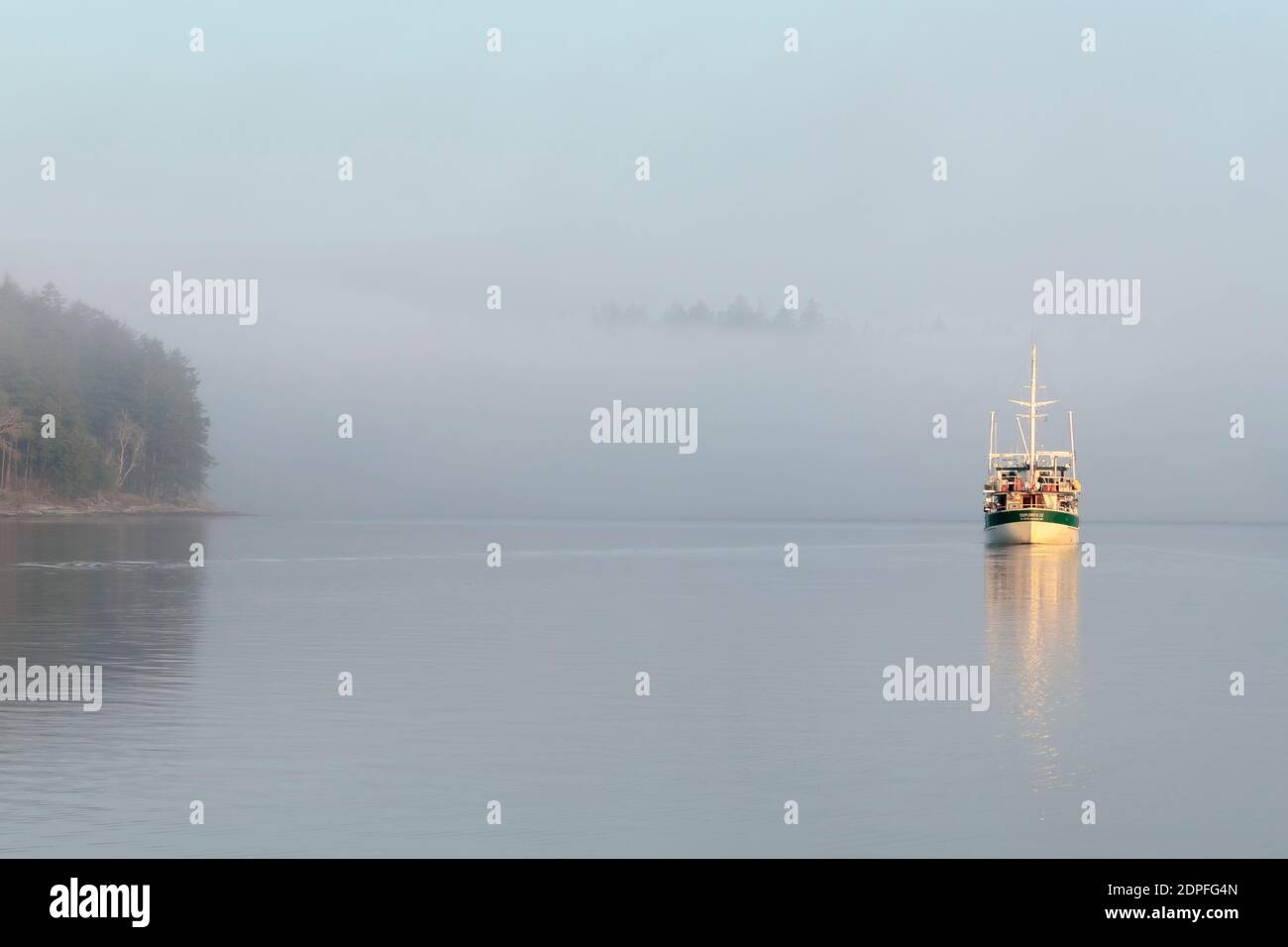 Un barco de placer está en marcha con cautela en una mañana niebla, no hay monumentos visibles excepto un punto cercano y unas cuantas copas de los árboles en una costa distante. Foto de stock