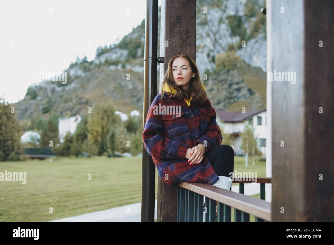Mujer joven en abrigo sentada en cercado de madera cerca de la cabaña en  las montañas de fondo Fotografía de stock - Alamy