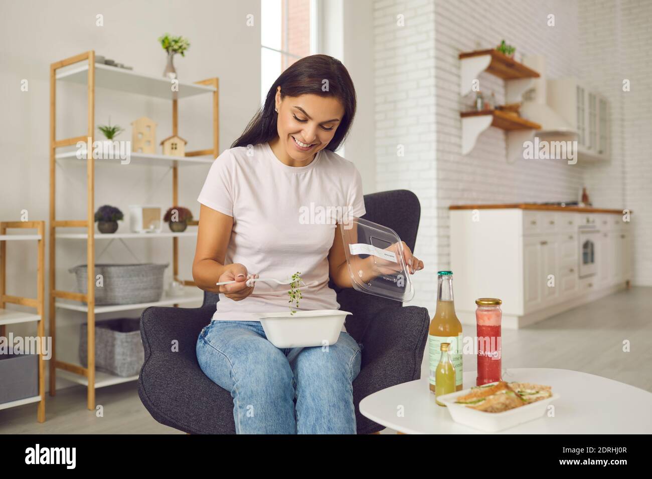 Feliz mujer joven con un almuerzo fresco y saludable para llevar sentado un sillón en casa Foto de stock