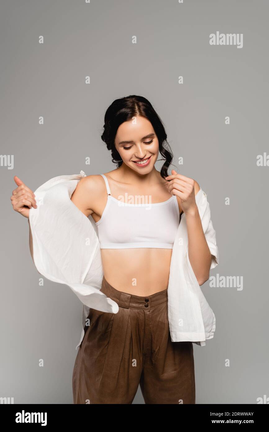 Mujer morena sonriente en la parte superior y pantalones de cuero,  quitándose camisa blanca aislada en gris Fotografía de stock - Alamy