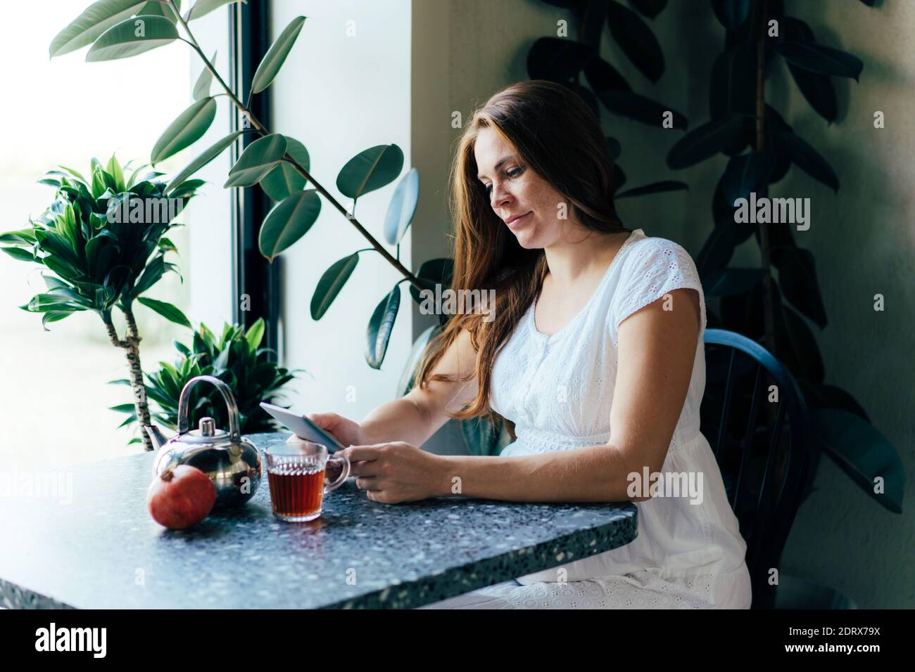 Una mujer embarazada encantadora está leyendo un libro electrónico en una mesa en un café. Foto de stock