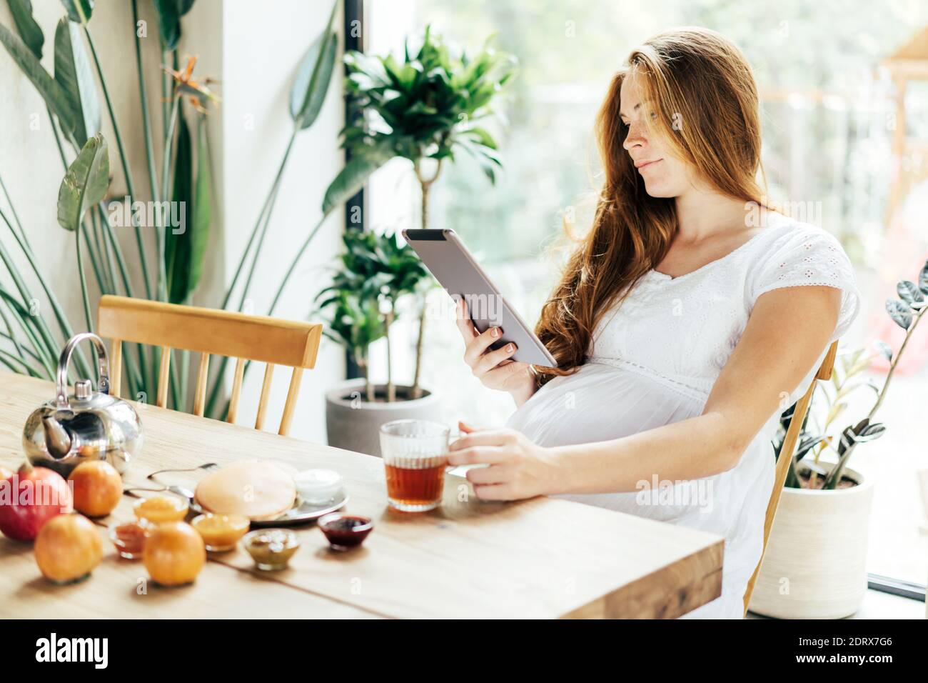 Una mujer embarazada está desayunando en la mesa y mirando en una tableta. Foto de stock