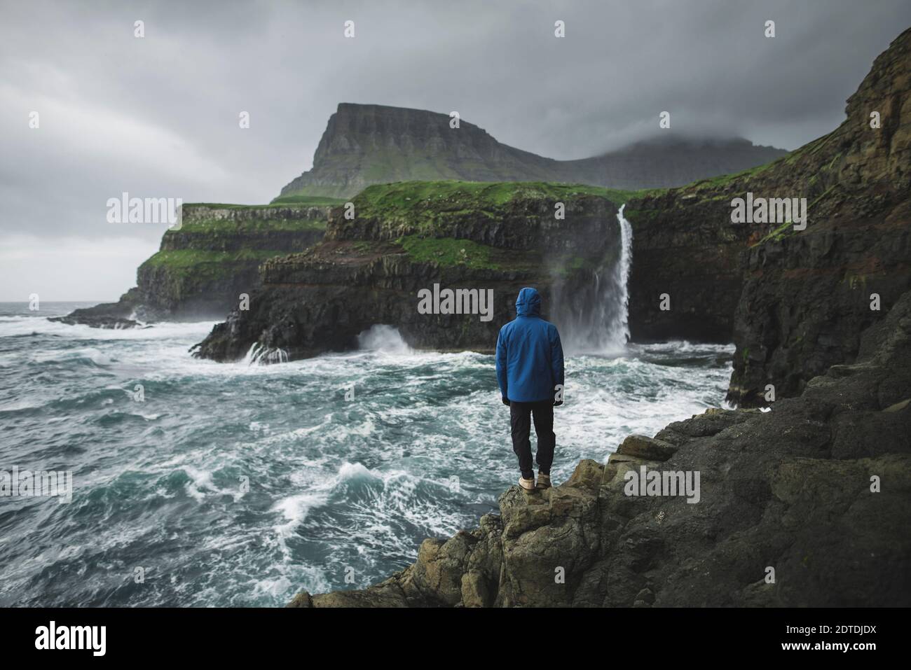 Dinamarca, Islas Feroe, Pueblo Gasadalur, Cascada M·Lafossur, Hombre de pie en el acantilado y mirando el día de la Cascada Mulafossur durante la tormenta Foto de stock