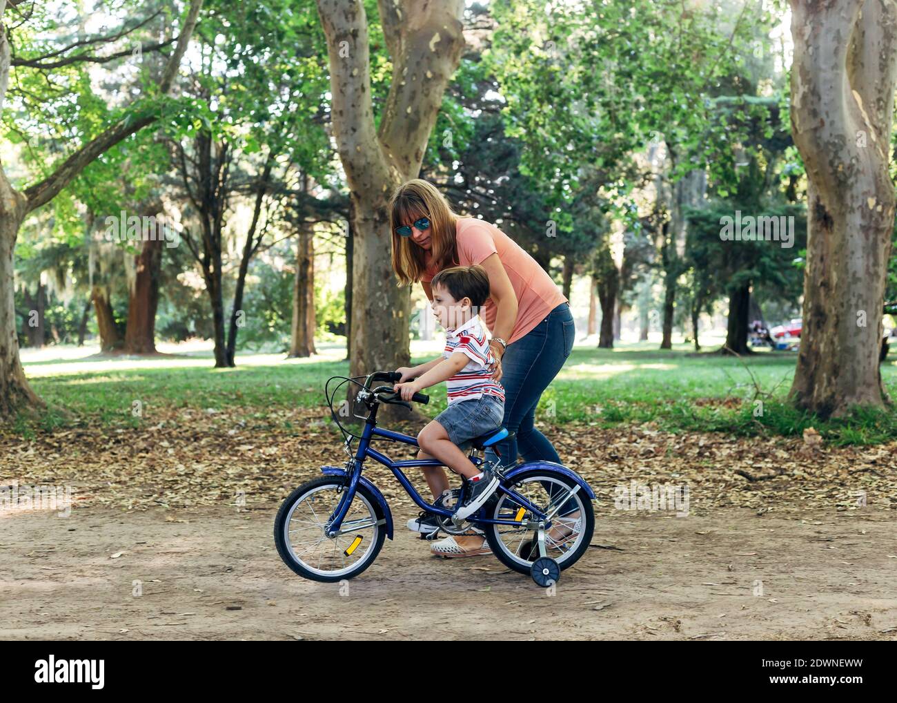 Mamá enseña a su hijo pequeño a montar en bicicleta. Un niño pequeño montando una bicicleta azul en una carretera sucia en un parque forestal y ayudado por su madre. Bicicleta con th Foto de stock