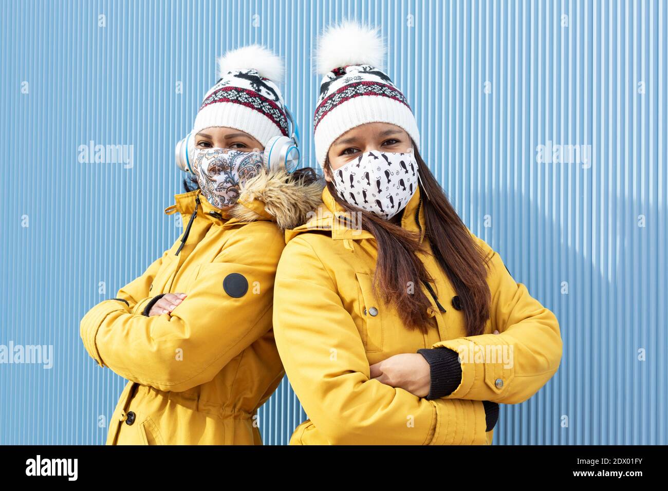 Las jóvenes parejas con máscaras faciales y ropa de invierno haciendo los brazos cruzados gesto. Están aislados sobre fondo azul. Conc. Protección contra coronavirus Foto de stock