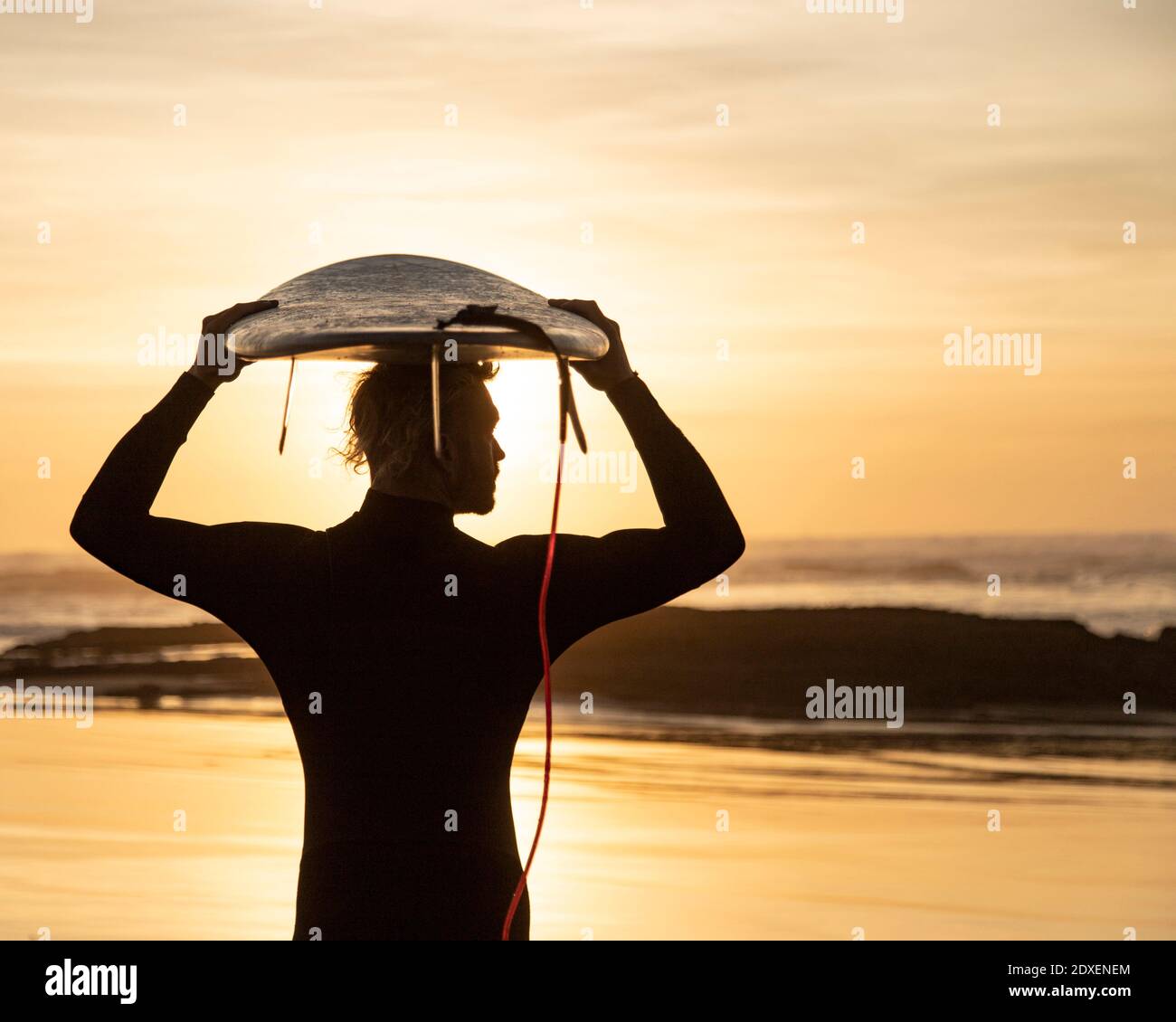 Surfista silueta llevando tabla de surf sobre la cabeza en la playa durante la puesta de sol Foto de stock