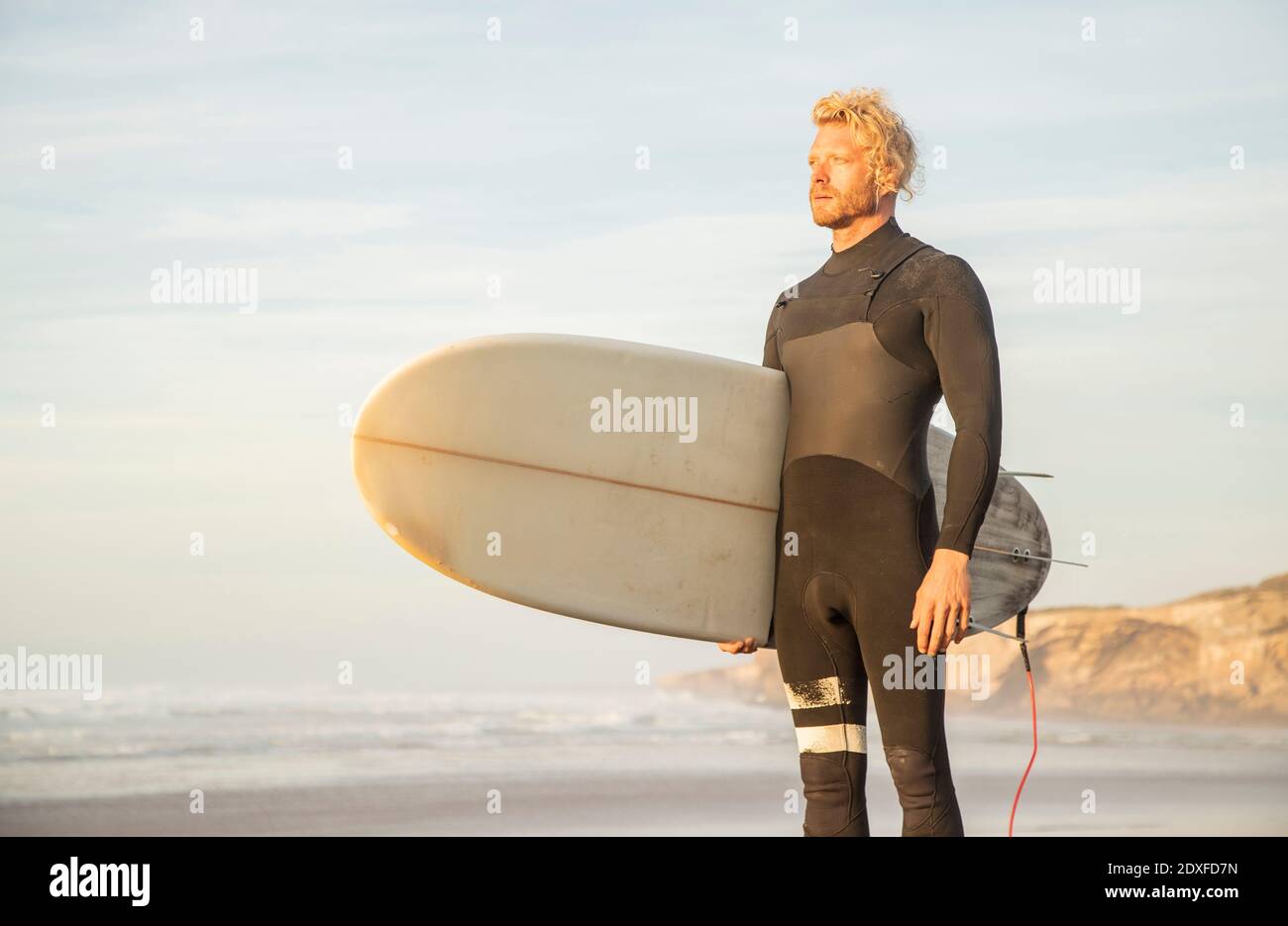 Hombre rubio con traje de neopreno llevando tabla de surf mientras mira lejos playa contra el cielo Foto de stock
