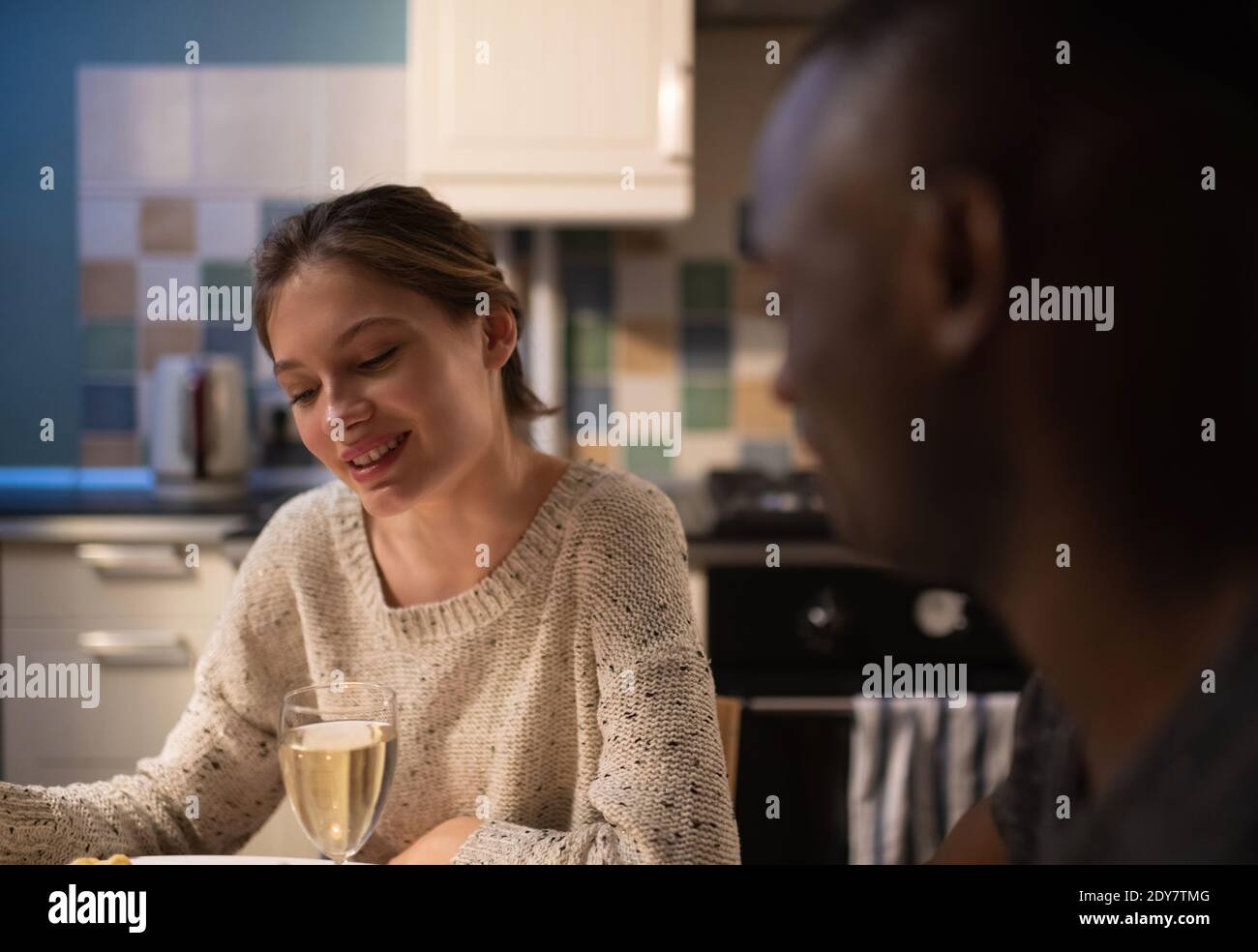 Mujer joven con una copa de vino sonriendo y hablando con Borroso afroamericano durante la cena romántica en casa Foto de stock