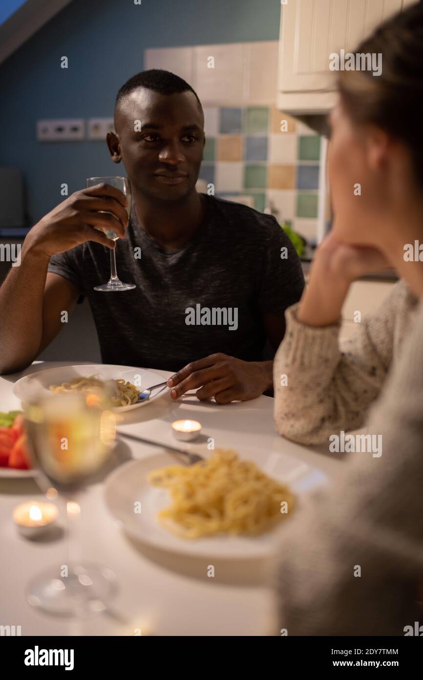 Hombre afroamericano con copa de vino escuchando borroso novia mientras se sienta en la mesa de la cocina durante la cena romántica en inicio Foto de stock