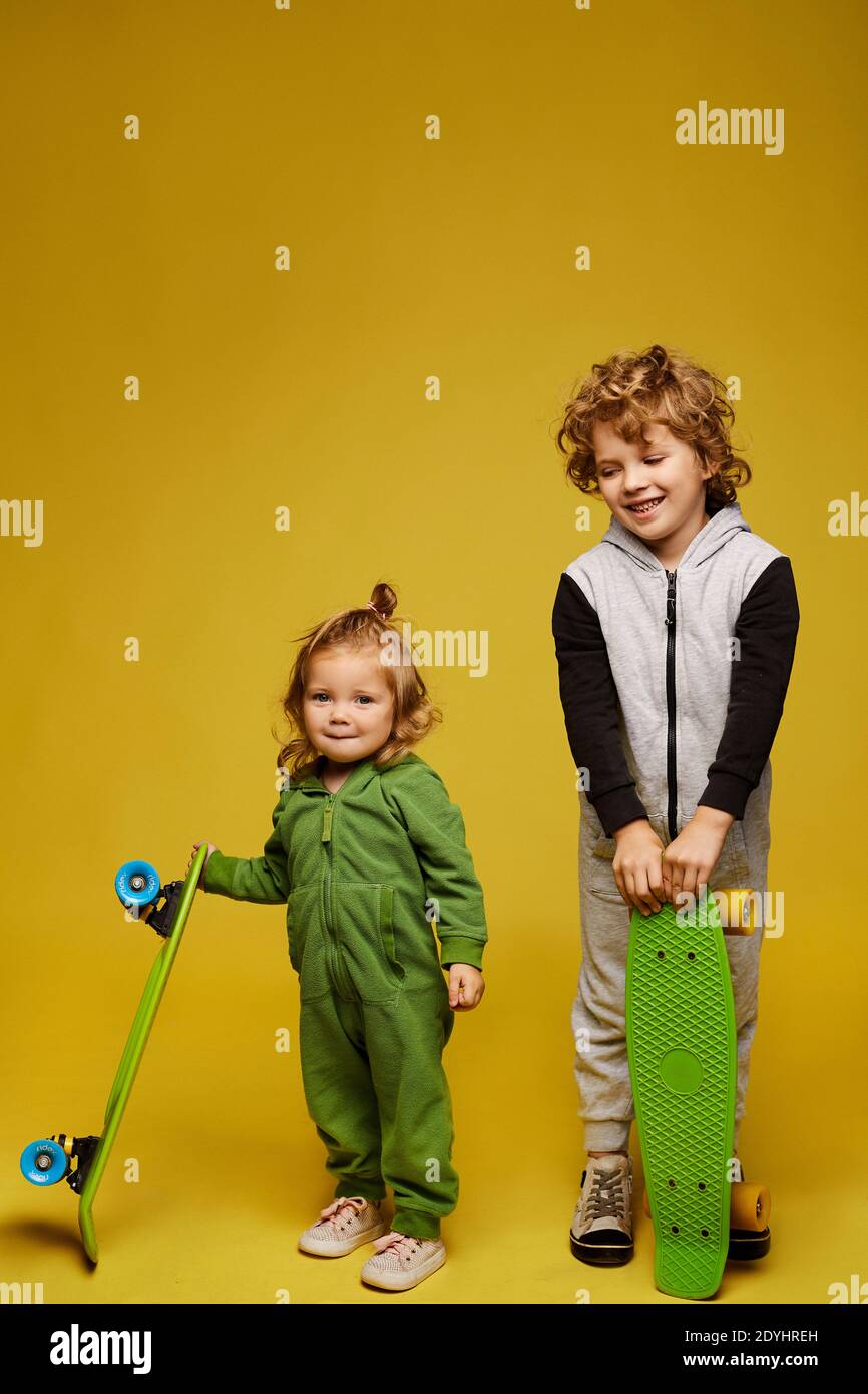 Feliz niño rizado y linda niña en kigurumi sosteniendo longboards y posando sobre el fondo amarillo, aislado. Dos niños sonriendo en trajes de verano Foto de stock