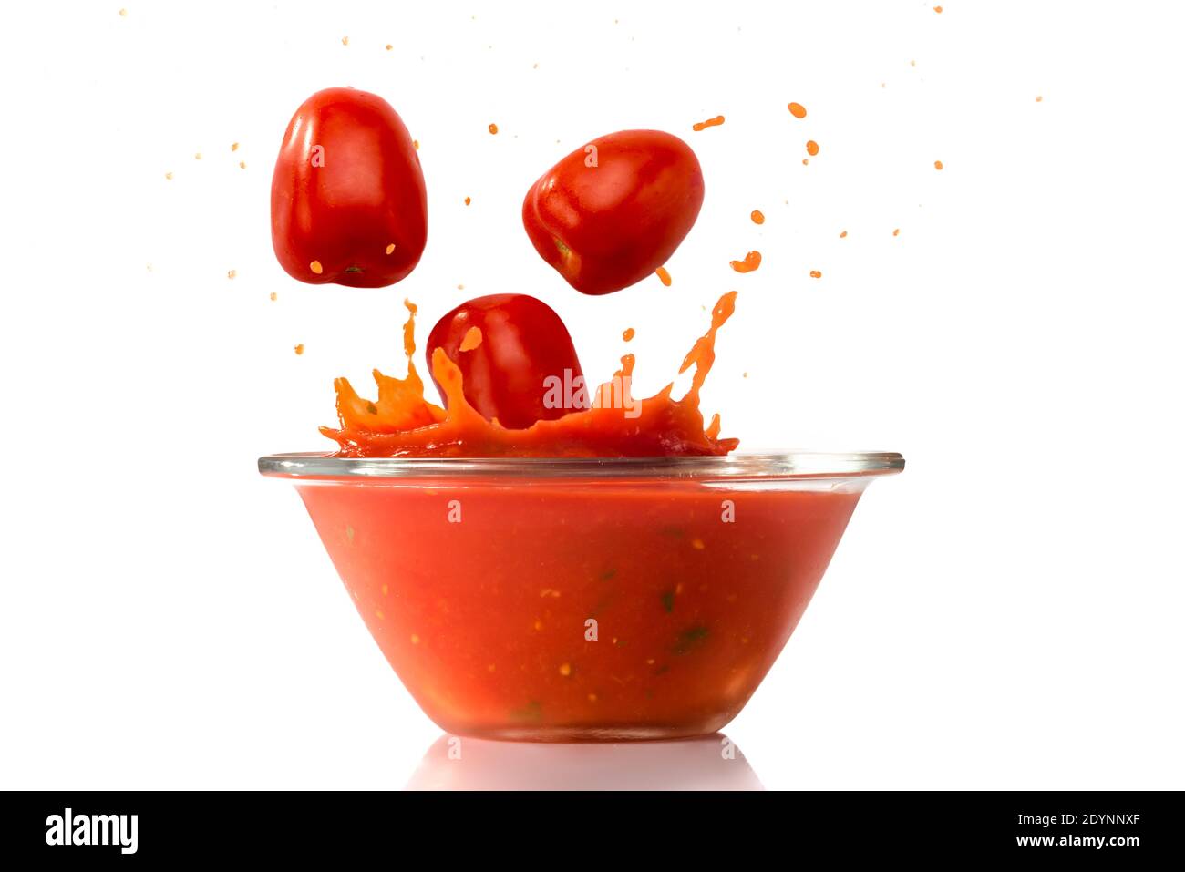 Los tomates maduros caen en un tazón de vidrio con salsa de tomate rojo salpicado, aislado sobre fondo blanco Foto de stock