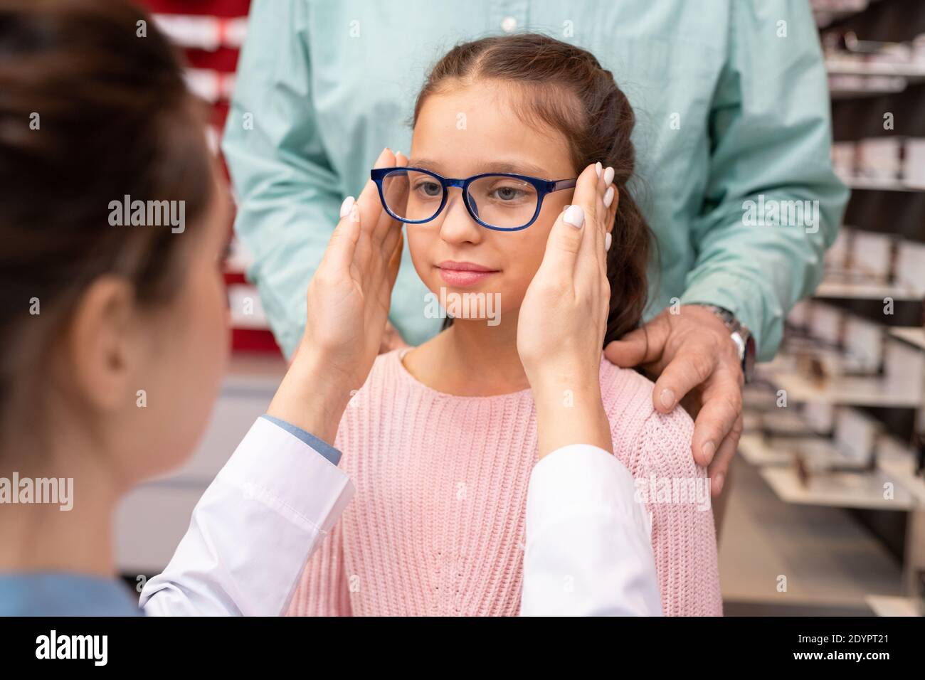 Joven oftalmóloga femenina en witecoat poniendo un par de gafas nuevas en la cara de la pequeña muchacha linda que visita la oficina médica con su abuelo Foto de stock