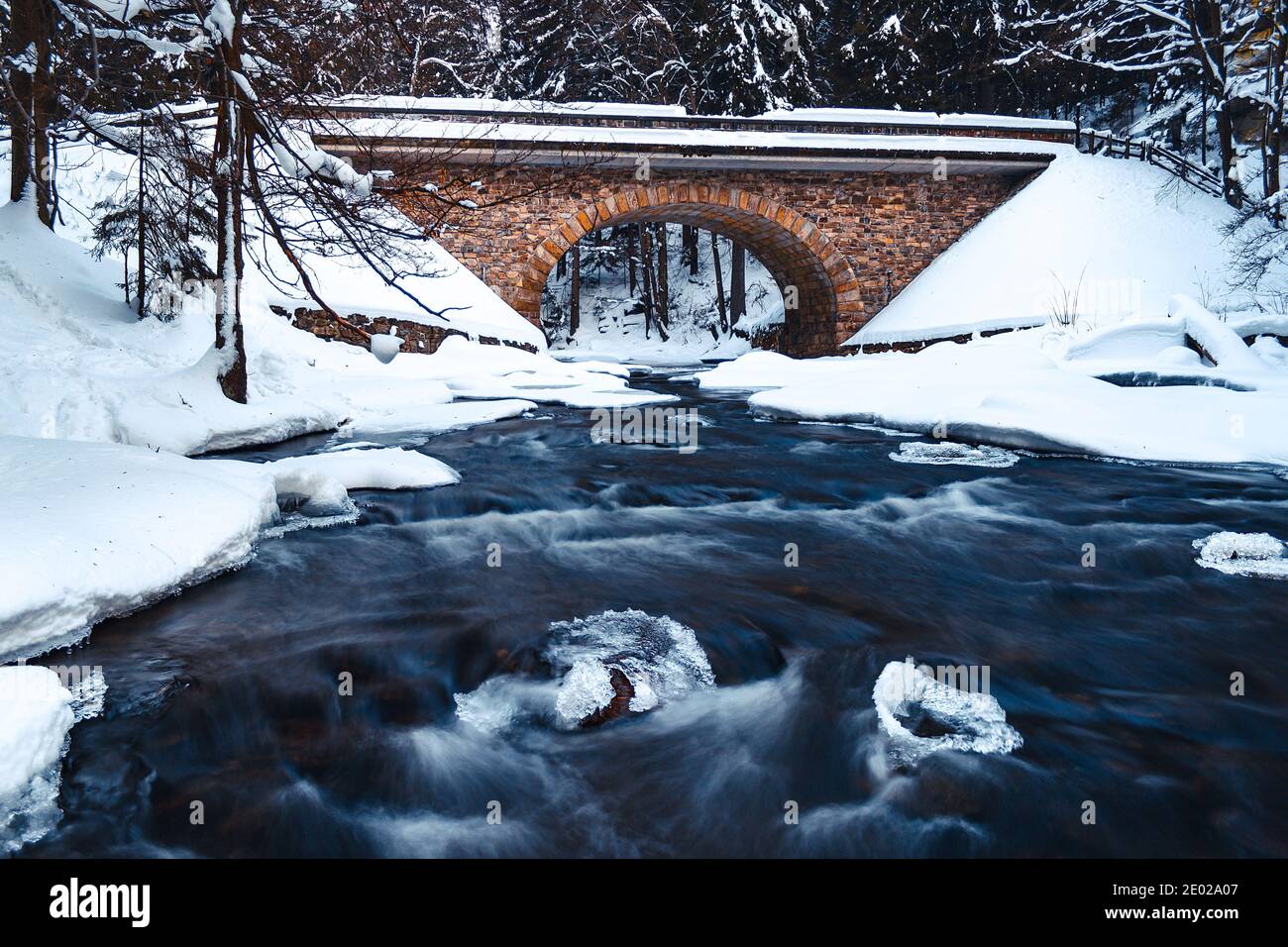 Viejo puente de piedra sobre el río salvaje en invierno, rodeado de bosques, nieve y hielo. El agua es borrosa por una exposición larga. Foto de stock