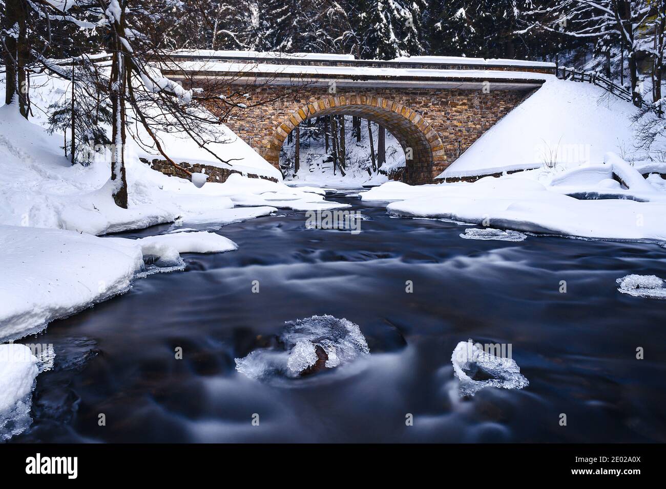Viejo puente de piedra sobre el río salvaje en invierno, rodeado de bosques, nieve y hielo. El agua es borrosa por una exposición larga. Foto de stock