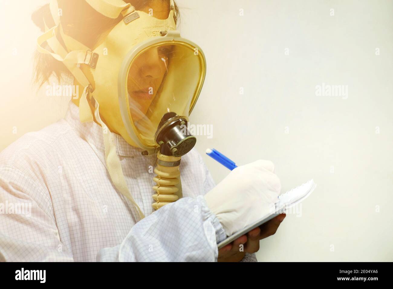 Una mujer con una máscara de oxígeno protectora de cara completa, abrigo blanco y guantes de goma, preparándose para trabajar en un laboratorio bioquímico, tomando notas con su pluma A. Foto de stock