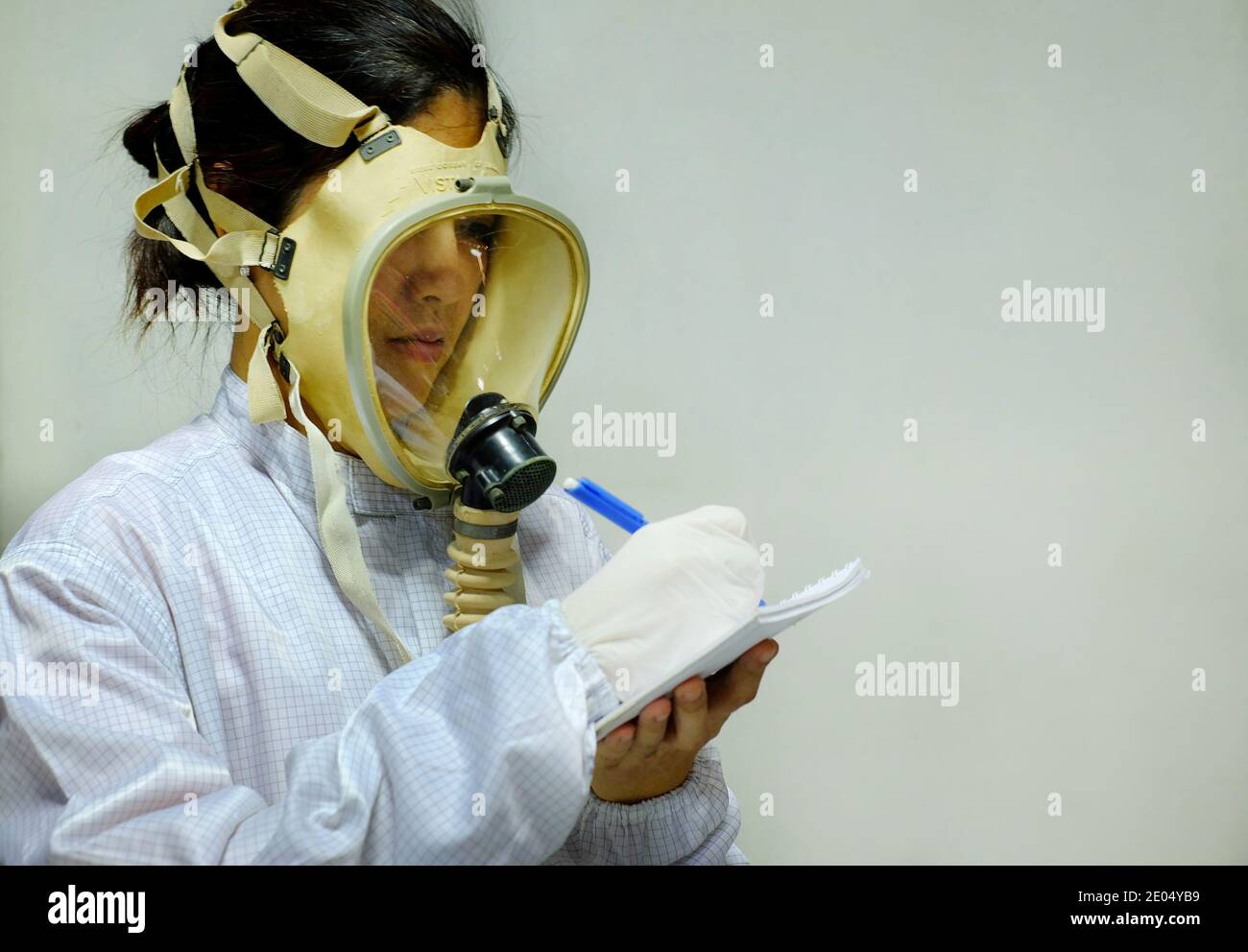Una mujer con una máscara de oxígeno protectora de cara completa, abrigo  blanco y guantes de goma, preparándose para trabajar en un laboratorio  bioquímico, tomando notas con su pluma A Fotografía de