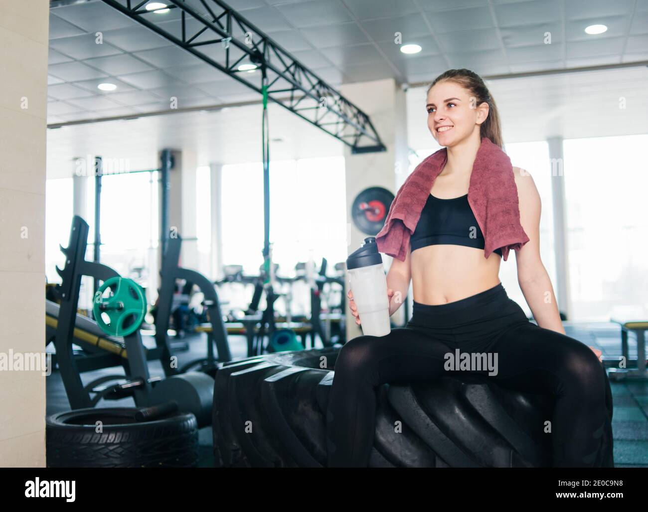 Mujer joven en sportswear con toalla alrededor de su cuello sosteniendo  botella de agua y sentado en la rueda grande para entrenamiento funcional  en el gimnasio Fotografía de stock - Alamy