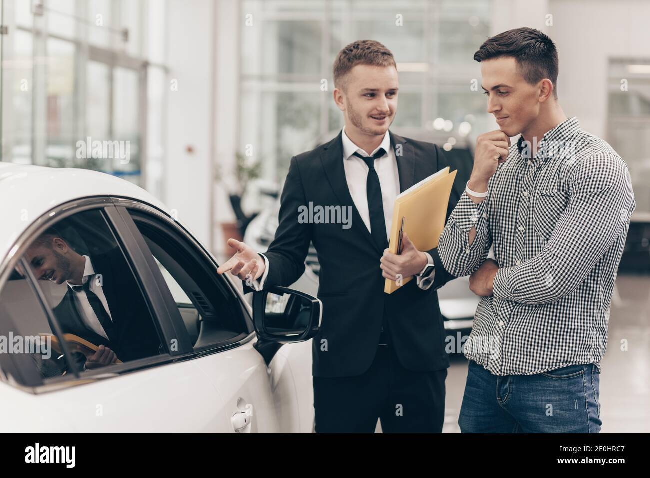 Concesionario profesional de coches mostrando coches a la venta a su cliente masculino, trabajando en el salón del concesionario de automóviles. Hombre mirando el coche con atención, es una exa Foto de stock