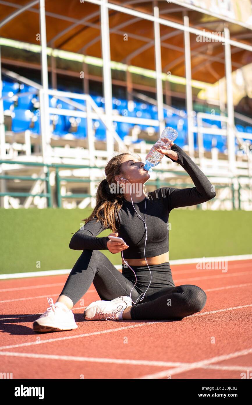 Mujer joven sedienta en forma bebiendo agua de la botella de plástico, descansando después de correr en un estadio de goma de la rueda de ardilla en el soleado día de verano, sosteniendo el movimiento sm Foto de stock
