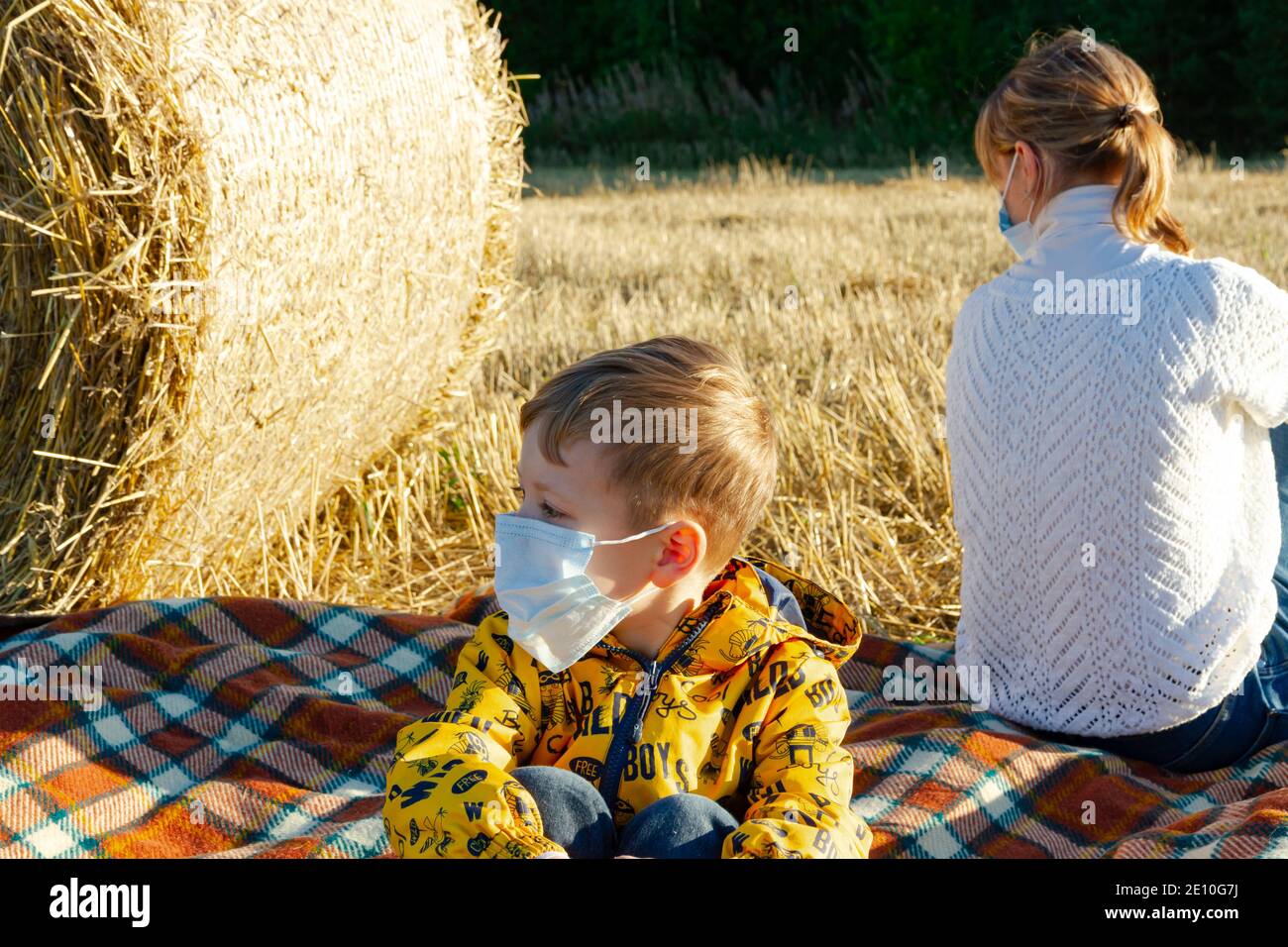 mamá y el bebé se sientan en el suelo en la naturaleza el fondo de los pajar en máscaras médicas Foto de stock
