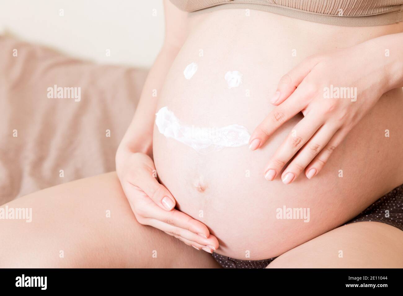 una niña embarazada se sienta en casa en la cama y se mancha con una crema antiestrías en el estómago. Embarazo, maternidad, preparación y expectativa Foto de stock