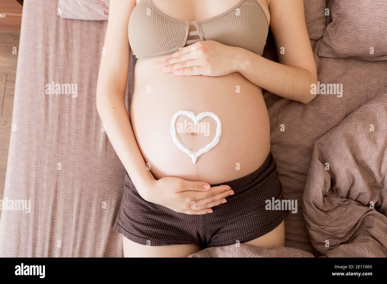 una niña embarazada se sienta en casa en la cama y le mancha el corazón con una crema antiestrías en el estómago. Embarazo, maternidad, preparación y expectativa Foto de stock